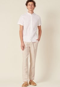 Camicetta bianca a maniche corte con bottoni in lino, abbinata a pantaloni in lino beige e scarpe slip-on color nocciola. Il modello è in piedi con una mano in tasca.
