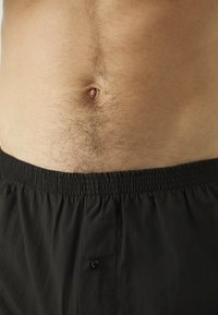 Shorts noirs à taille élastique avec un détail de bouton, portés sur une peau bronzée montrant des cheveux châtains clairs. Mettez l'accent sur la texture du tissu et l'ajustement.