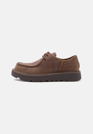 Scarpa marrone in pelle con lacci, suola spessa e scura e cuciture in stile mocassino, mostrata di lato su sfondo bianco.