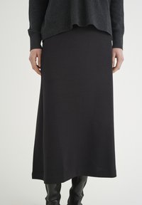 Černá jersey sukně po kotníky s hladkou texturou a uvolněným střihem, spárovaná s tmavým, splývavým topem. Silueta je minimalistická.