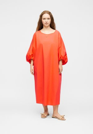 Naine seisab, kandes lahtist, erkoranži midi-kleiti koos volangiliste varrukatega ja beeže sandaale ühetaustalise valge taustaga.