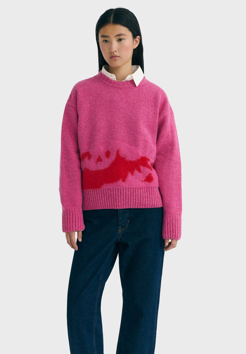 Ung kvinde bærer en pink og rød tekstureret sweater over en hvid skjorte med krave og løse mørkeblå jeans foran en ensfarvet baggrund.