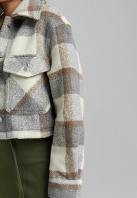 Krótka kurtka flanelowa w kratę w odcieniach szarości, bieli i brązu. Posiada dwie przednie kieszenie, zapięcie na guziki oraz miękką teksturę.