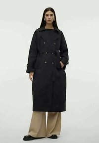 Czarny płaszcz trench z podwójnym zapięciem i paskiem, z szerokimi klapami i dwoma bocznymi kieszeniami. Modelka zestawia go z beżowymi spodniami o szerokich nogawkach.