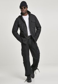 Black cargo jacket with two chest pockets, paired with loose-fitting black cargo pants. Worn with a white t-shirt, black beanie, and sunglasses.