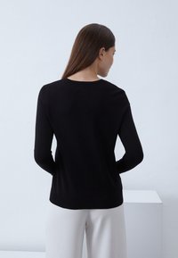 Maglione nero a maniche lunghe con scollatura rotonda, texture liscia e design aderente. La schiena del modello è visibile, abbinata a pantaloni chiari.