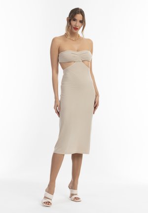 Frau mit zurückgebundenem Haar, die ein beiges trägerloses Midi-Kleid mit Ausschnitten, weiße Sandalen mit Absatz und eine auffällige goldene Halskette trägt, stehend vor weißem Hintergrund.
