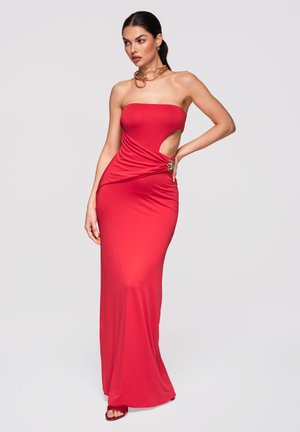Kobieta stojąca w czerwonej, długiej do ziemi sukni bez ramiączek z bocznym wycięciem i marszczonym detalem, nosząca złoty naszyjnik typu choker i czerwone sandały.