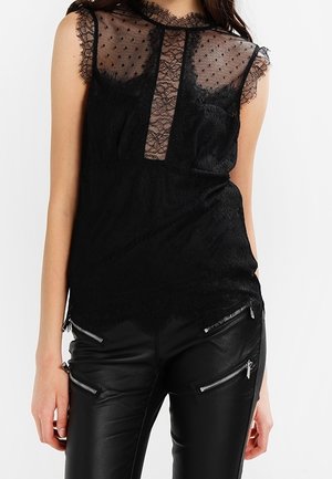 Top nero senza maniche con un corpetto in rete trasparente a pois e dettagli in pizzo, abbinato a pantaloni in pelle nera con accessori con zip.
