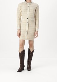 Robe en tricot crème et beige à motif à carreaux, avec des boutons, deux poches avant et des manches longues ; associée à des bottes de cowboy hautes, marron foncé.
