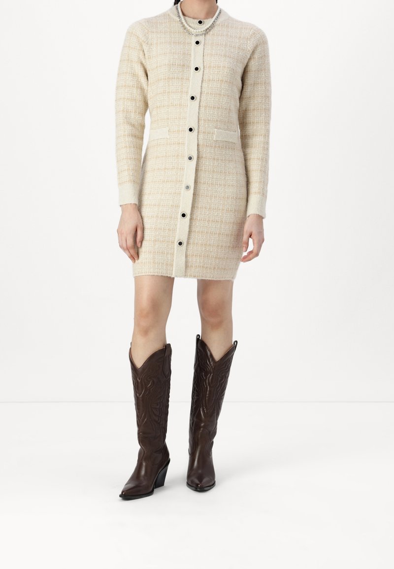 Robe en tricot crème et beige à motif à carreaux, avec des boutons, deux poches avant et des manches longues ; associée à des bottes de cowboy hautes, marron foncé.