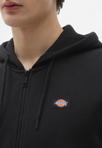 Sort zip-up hoodie med snore, tekstureret stof og et farverigt "Dickies" logo patch på venstre bryst.