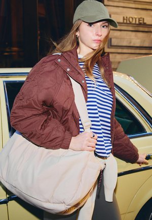 Tânără purtând o șapcă verde, o cămașă cu dungi și o jachetă maro, ținând o geantă mare bej în timp ce deschide portiera unei mașini galbene lângă un hotel.