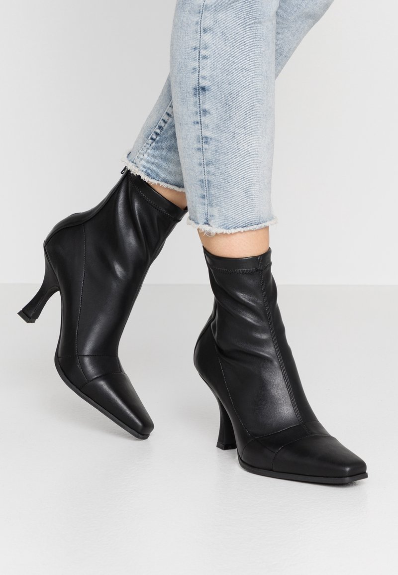 Bottines à talons en cuir noir à bout pointu portées avec un jean à ourlet effiloché délavé clair sur un fond blanc uni.
