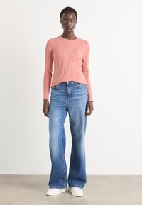 Pulover tricotat cu model cablu, de culoare roz pal, asortat cu blugi evazați din denim albastru. Blugii au un gradient decolorat și un design clasic cu cinci buzunare.