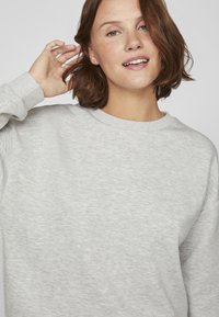 Joven con cabello corto castaño que sonríe, vestida con un sudadera de cuello redondo gris claro, con la mano levantada cerca de su oído frente a un fondo liso.