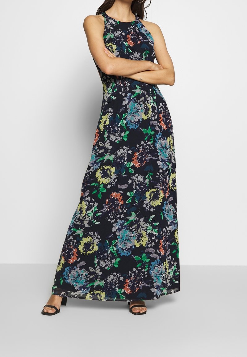 Femme debout, les bras croisés, portant une robe noire sans manches longue jusqu'au sol avec des motifs floraux multicolores et des talons noirs ouverts à l'avant.