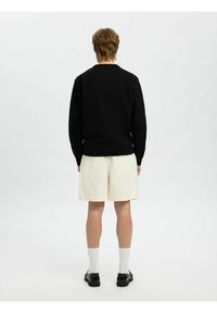 Sudadera de algodón negra con puños acanalados, combinada con pantalones cortos crema. El modelo lleva calcetines blancos y zapatos negros. Fondo neutro.