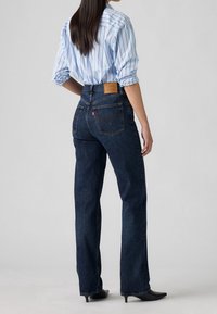 Kvinde stående med ryggen mod, iført mørkeblå højtaljede lige jeans, lyseblå stribet skjorte med op rullede ærmer og sorte spidse sko.