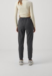 Kvinna iklädd en krämfärgad långärmad tröja och mörkgrå sweatpants med elastisk midja samt sneakers, stående vänd bort från kameran på en enfärgad bakgrund.