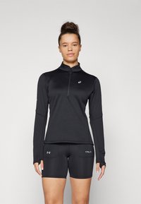 Maglia sportiva nera a maniche lunghe con collo zip e tessuto strutturato, abbinata a pantaloni corti sportivi neri con tasche laterali e marchio.