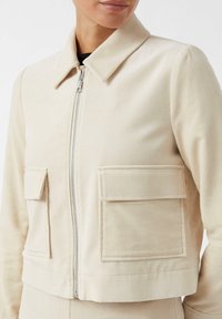 Beige corduroyjack met een kraag, een ritssluiting aan de voorkant en twee vierkante zakken op de borst, met een cropped pasvorm en geribbelde textuur.