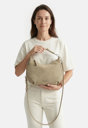 Vrouw in witte outfit met een beige schoudertas met ritssluiting en een lange verstelbare band tegen een effen achtergrond.