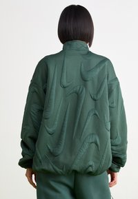 Προστατευτικό πράσινο μπουφάν με ανάγλυφα σχέδια Nike swoosh, ψηλό γιακά και χαλαρή εφαρμογή με ελαστικές μανσέτες και στρίφωμα.