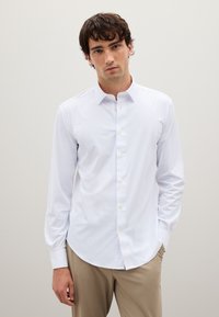 Camisa blanca de manga larga con botones, hecha de una tela suave; presenta un cuello clásico y detalles de botones en la parte delantera. Llevada con pantalones beige.