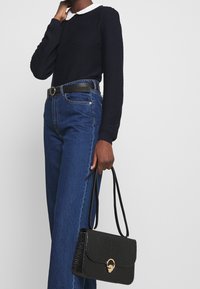 Svart väskan i konstläder med krokodilstruktur och ett spänne i guldmetall, hålls i en hand; matchas med en mörk tröja och blå jeans med hög midja.