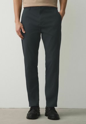 Ein Mann trägt schwarze gerade geschnittene Hosen und schwarze Schnürschuhe und steht vor einem einfarbigen hellen Hintergrund.