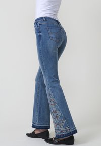 Jeans de mezclilla azul acampanados con intrincados bordados multicolores en el dobladillo y la costura lateral, con detalles de bolsillos tradicionales.