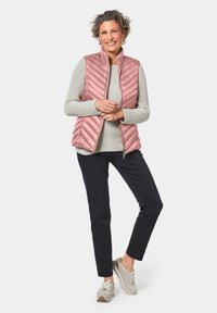 Lichtroze gewatteerde vest over een grijze trui, in combinatie met donkere broek en lichte sneakers. Het vest heeft een opstaande kraag en een ritssluiting.