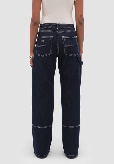 Personne portant un jean à jambes larges bleu foncé avec des surpiqûres blanches contrastées, des poches plaquées et une boucle pour outil, vue de dos sur un fond uni.