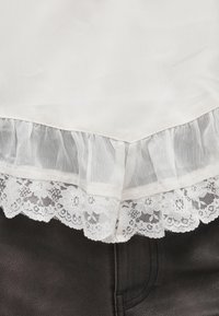 Biela látka s priehľadným volánom a čipkovou lemovkou na tmavom textúrovanom koženom materiáli, zobrazujúca vrstvenie a textúry odevu.