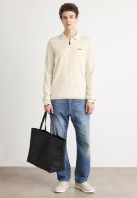 Long-sleeve cream polo with a zip collar, black logo, paired with blue jeans and beige sneakers, holding a large black tote bag.