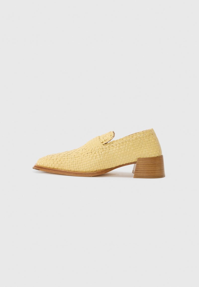 Sapatilha em couro amarelo trançado com bico fino, salto baixo de madeira e uma pequena aba decorativa na parte superior. Detalhe de superfície texturizada.
