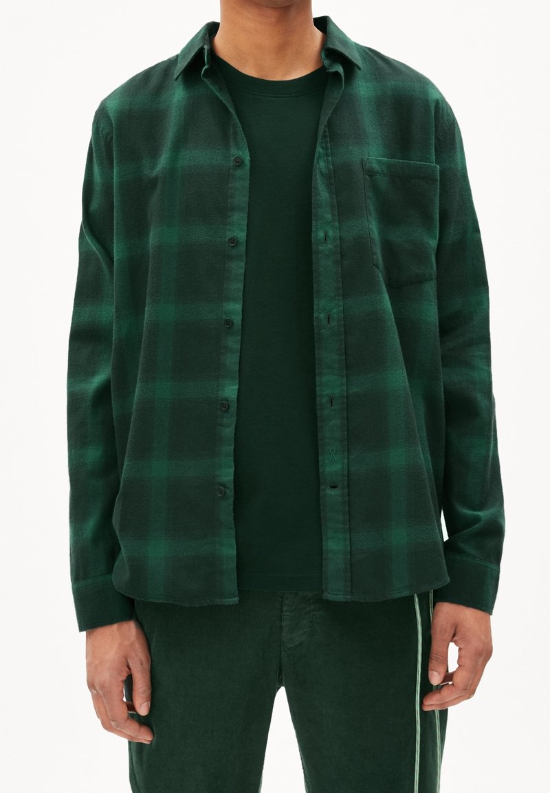 Uomo che indossa una camicia a quadri verde scuro sbottonata sopra una maglietta verde scuro uniforme e pantaloni verde scuro abbinati con striscia laterale.