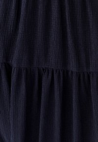 Tumši zils kropls audums ar teksturētu virsmu, kas veidots ar kārtainu dizainu un savāktām šuvēm, radot mīkstu un vieglu izskatu.