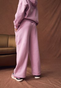 Sudadera con capucha rosa claro y pantalones anchos a juego hechos de tela suave, con bolsillos profundos y un ajuste cómodo. El calzado que se muestra es multicolor.