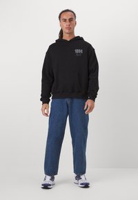 Sweatshirt à capuche noire avec poche avant, orné d'un design imprimé gris. Porté avec un jean en denim bleu et des baskets multicolores.