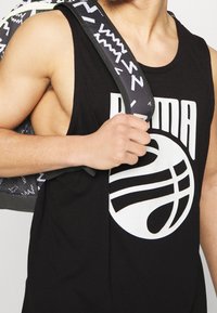 Homme portant un débardeur noir avec un logo de basket-ball blanc, portant un sac à dos noir et blanc à motifs sur une épaule.