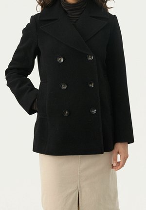 Femme portant un manteau en laine noir à double boutonnage sur un col roulé rayé, associé à une jupe beige avec une fente à l'avant.