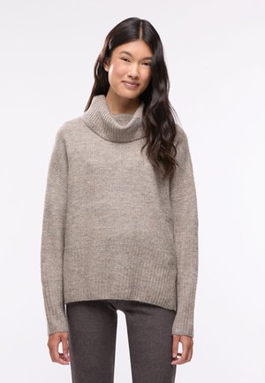 Donna con lunghi capelli scuri che indossa un maglione dolcevita beige e pantaloni grigio scuro, in piedi su uno sfondo chiaro e uniforme.