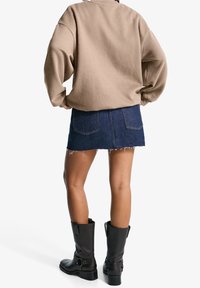 Osoba stojąca z rękami na biodrach, ubrana w beżową bluzkę z długim rękawem, ciemnoniebieską dżinsową mini spódniczkę oraz czarne kozaki do połowy łydki na białym tle.
