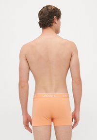 Man gezien van achteren in oranje Lacoste boxershorts tegen een effen witte achtergrond.