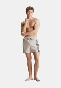 Pantaloncini da bagno beige con cordoncino nero e stampa del logo laterale. Presentano una vita elastica, lunghezza fino a metà coscia e una texture liscia.