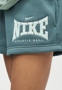 Κοντινή λήψη από τυρκουάζ σορτς Nike με κεντημένο λευκό λογότυπο και κείμενο "Athletic Dept." στο πάνω μέρος του μηρού ενός ατόμου.