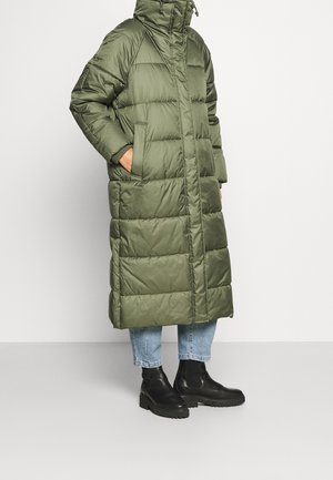 Długa oliwkowozielona pikowana kurtka puchowa z kapturem, przednimi kieszeniami, noszona na skróconych jasnoniebieskich dżinsach i czarnych botkach za kostkę.