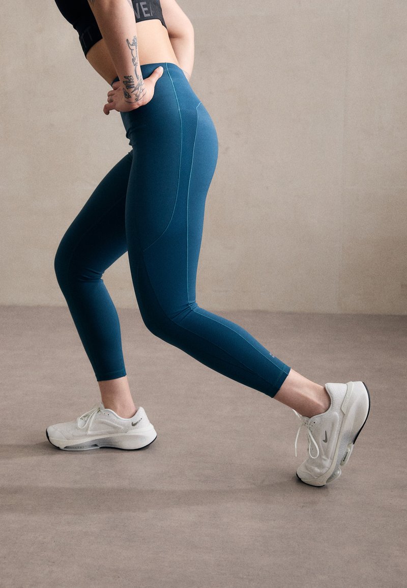 Leggings atletici turchesi con una texture liscia e cuciture a contrasto, abbinati a sneaker bianche con un logo discreto e dettagli di ammortizzazione.
